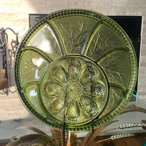 May include: A vintage green glass serving platter with a floral and leaf design. The platter is divided into sections, with a central flower-shaped area and surrounding compartments. The rim has a textured, beaded edge. A decorative piece for serving appetizers or snacks.