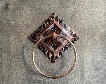 May include: Ornate bronze-colored towel ring with a decorative diamond-shaped backplate. The ring is circular and made of a gold-colored metal. The backplate features intricate scrollwork and is mounted on a wooden surface.