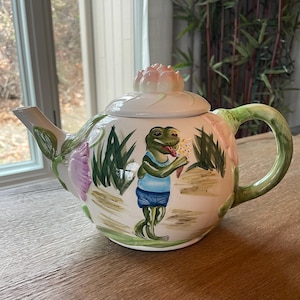 May include: A white ceramic teapot with a green frog design. The frog is wearing a blue shirt and holding a flower. The teapot has a pink flower on the lid.