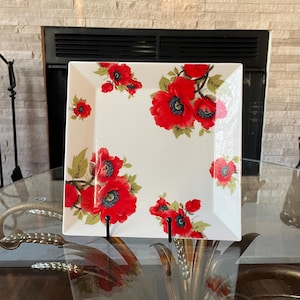 May include: A square, white ceramic serving platter with a vibrant floral design. The pattern features large, red poppy flowers with black centers and green leaves. The platter is displayed on a glass table.