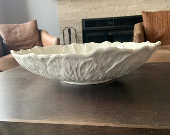May include: A large white ceramic bowl with a textured, cabbage-like design. The bowl is sitting on a wooden table.