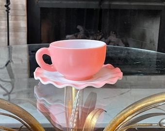 May include: A pink teacup and saucer set. The cup is a solid pink color with a curved handle. The saucer is white with a pink ruffled edge. The set is sitting on a glass table with a gold-colored base.