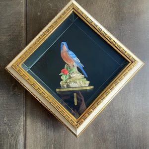 May include: A framed, diamond-shaped mirror with a decorative bluebird figurine. The bird has blue and orange plumage and sits on a small, detailed base with a red flower. The frame is white with gold accents.