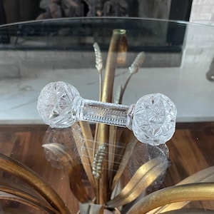 May include: A clear glass knife rest featuring two spherical ends with a faceted starburst pattern and a central cylindrical bar with a textured, beaded surface. The object is transparent and reflects light through its intricate geometric cut glass design.