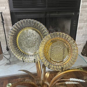 May include: Two decorative glass plates with intricate embossed patterns. One plate is clear with a gold rim, and the other is amber-colored. The plates are displayed on a glass table with a gold metal base, showcasing a vintage aesthetic.