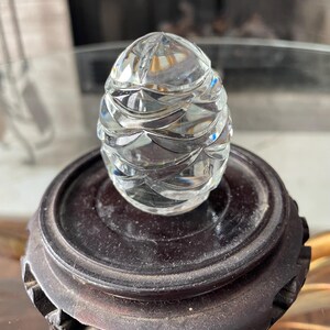 May include: A clear, faceted glass egg-shaped object sits atop a dark brown wooden stand. The egg has a detailed, textured surface that reflects light. The stand is round with a carved design, showcasing a vintage decorative piece.