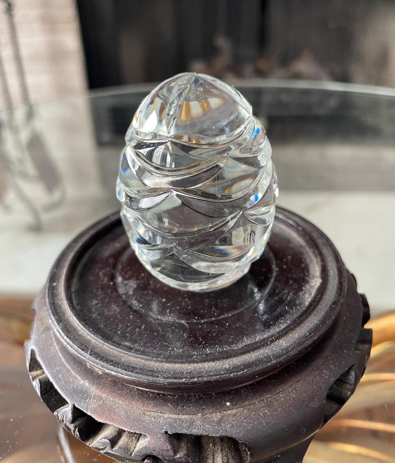 May include: A clear, faceted glass egg-shaped object sits atop a dark brown wooden stand. The egg has a detailed, textured surface that reflects light. The stand is round with a carved design, showcasing a vintage decorative piece.