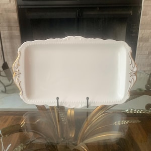 May include: A white rectangular serving tray with a scalloped edge and gold accents. The tray is supported by a black metal stand and is displayed on a glass surface. The tray has a decorative design.