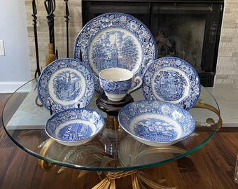 May include: A collection of blue and white ceramic tableware, including plates, bowls, and a teacup. The pieces feature a detailed landscape design. The set is arranged on a glass-topped table, showcasing the classic aesthetic of the dinnerware.