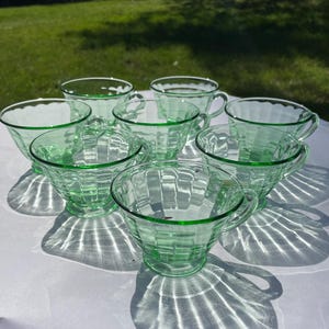 May include: A set of eight green glass teacups. Each cup has a ribbed design and a handle. The cups are arranged on a white surface, with shadows cast by the sunlight. The background is a blurred green lawn.
