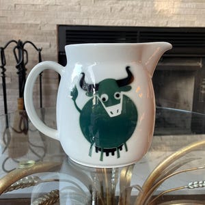 May include: White ceramic pitcher with a green cartoon bull design. The pitcher has a handle and a spout. The bull has black horns and a white face. The pitcher is sitting on a glass table.