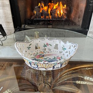 May include: A white porcelain openwork bowl with a colorful pheasant and floral design. The bowl has a scalloped edge and is sitting on a glass table. The background features a fireplace with a burning fire.