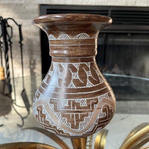 May include: A brown ceramic vase with a wide mouth and a bulbous body. The vase features intricate white carved designs, including geometric patterns and leaf-like shapes. The vase is displayed on a glass table.