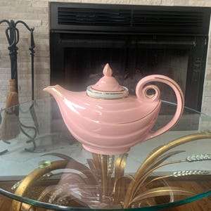 May include: A pink ceramic teapot with a lid, featuring gold trim and a curved handle. The teapot is displayed on a glass table with a decorative gold base, set against a neutral background.