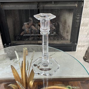 May include: A clear, faceted crystal candlestick holder with a hexagonal base and top. The candlestick has a tall, slender stem and is designed to hold a single candle. The holder is displayed on a glass table with decorative gold accents.