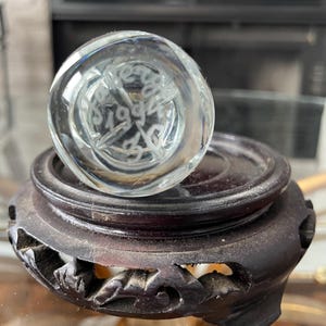 May include: A clear glass paperweight with etched text and a starburst design, resting on a dark brown, carved wooden stand. The paperweight has the etched text "1994". The stand has a round top and carved floral designs.