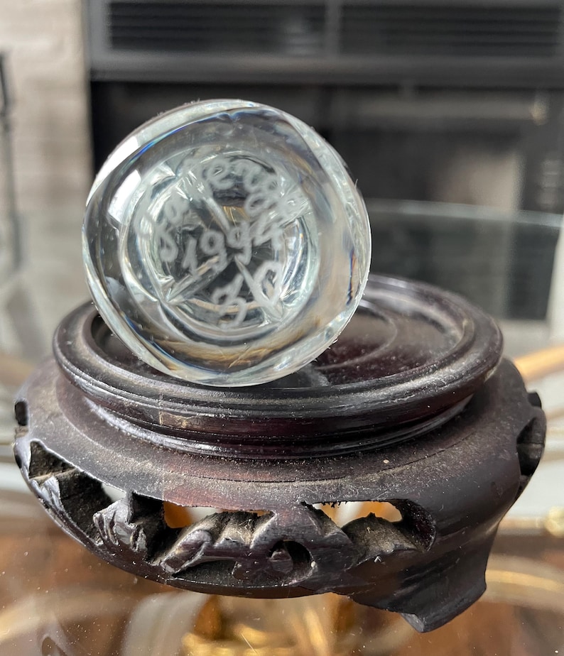 May include: A clear glass paperweight with etched text and a starburst design, resting on a dark brown, carved wooden stand. The paperweight has the etched text "1994". The stand has a round top and carved floral designs.