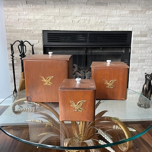 May include: Three wooden boxes of varying sizes with a brown finish and gold eagle emblem. Each box has a white knob on the lid. The boxes are displayed on a glass-topped table with a gold metal base.
