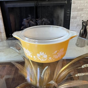 May include: A round, bright yellow casserole dish with a clear glass lid and integrated side handles. The exterior of the dish features a white floral pattern consisting of tulips and other small blossoms printed around the entire circumference.