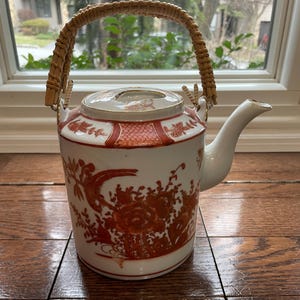 May include: White ceramic teapot with a woven handle and a curved spout. The teapot features red and gold floral and bird designs. The handle is made of woven light brown material. The teapot is a decorative item.
