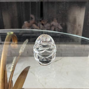 May include: A clear, faceted crystal egg-shaped decorative object. The egg has a series of horizontal, curved cuts that create a textured surface. The object is sitting on a glass surface, with a fireplace visible in the background.