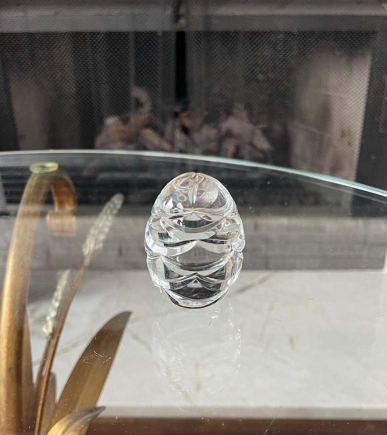 May include: A clear, faceted crystal egg-shaped decorative object. The egg has a series of horizontal, curved cuts that create a textured surface. The object is sitting on a glass surface, with a fireplace visible in the background.