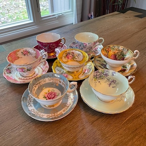 May include: A collection of vintage teacups and saucers in various colors and floral patterns. The teacups feature gold trim and intricate designs, including roses and other botanical motifs. The saucers complement the cups with matching patterns.