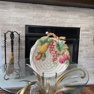 May include: A decorative ceramic fruit basket plate with a woven design, featuring colorful sculpted fruits like grapes, peaches, and figs. The plate is displayed on a glass-topped table with a gold metal base.