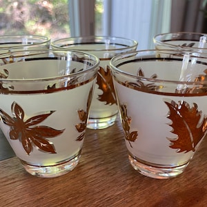 May include: Set of six vintage drinking glasses. Each glass features a white frosted background with gold leaf designs. The rims and leaf details are accented with gold. The glasses are arranged on a wooden surface.