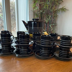 May include: A black tea set with white horizontal stripes, including a teapot, cups, and saucers. The set is arranged on a wooden table, with a window and greenery in the background. The cups are stacked.
