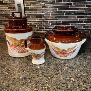 May include: A set of vintage ceramic kitchenware with a patriotic theme. The set includes a large canister, a small pitcher, and a covered pot. Each piece features a brown and white color scheme with an eagle and American flag design.