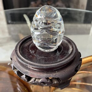 May include: A clear, faceted crystal egg sits atop a dark brown, carved wooden stand. The egg's surface reflects light, showcasing its intricate design. The stand has a circular top and a decorative base, adding to the piece's elegance.