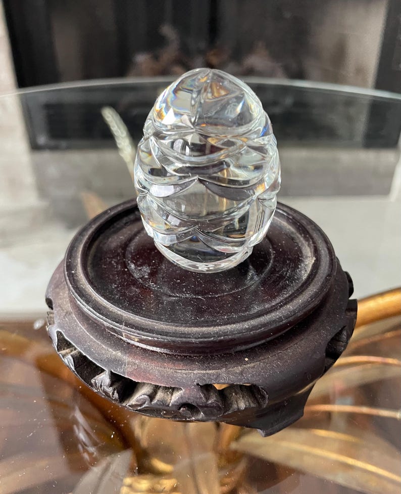 May include: A clear, faceted crystal egg sits atop a dark brown, carved wooden stand. The egg's surface reflects light, showcasing its intricate design. The stand has a circular top and a decorative base, adding to the piece's elegance.