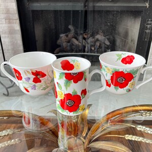 May include: Three white ceramic mugs with a floral poppy design. The mugs feature red poppies, yellow flowers, and green leaves. The mugs are on a glass table with a gold-colored decorative base.