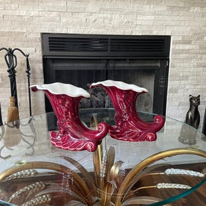 May include: Two decorative, red ceramic vases with a white interior, shaped like a horn of plenty. The vases are displayed on a glass-topped table with a gold-colored metal base, in front of a fireplace.