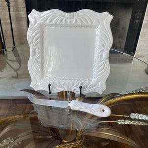 May include: A white, square, decorative serving plate with a detailed, ornate border. A matching white cheese knife rests in front of the plate. The plate and knife are displayed on a glass-topped table.