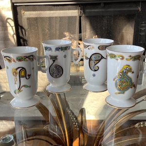 May include: Four white ceramic mugs with Celtic knot designs and gold rims. Each mug features a different letter or design in vibrant colors, including green, yellow, blue, and red. The mugs have a pedestal base and a handle.