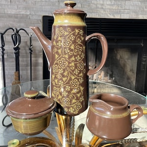 May include: A brown ceramic coffee set with a floral design. The set includes a tall coffee pot with a lid, a small creamer, and a covered sugar bowl. The coffee pot and sugar bowl have a gold accent around the rim.