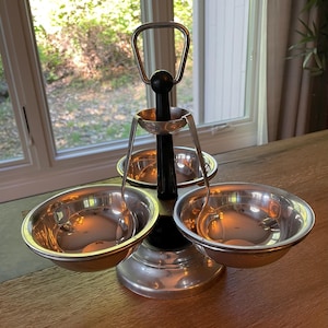 May include: A silver-toned serving dish with three bowls, a black central column, and a handle. The bowls are suspended by curved metal supports. The base is a decorative, tiered design. The background shows a window and a wooden table.