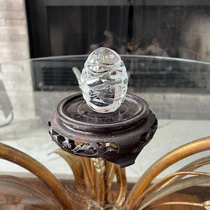 May include: A clear, faceted crystal egg sits atop a dark brown, intricately carved wooden stand. The stand is placed on a glass-topped table with a gold-colored, ornate base. The egg's facets catch the light, creating a sparkling effect.