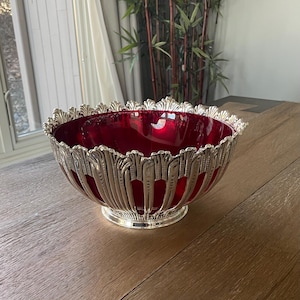 May include: A decorative bowl with a deep red interior and a silver-colored, ornate exterior. The bowl has a scalloped rim and a pedestal base. The exterior features a repeating, stylized leaf design. The bowl is sitting on a wooden surface.