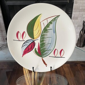 May include: A round white ceramic plate featuring a hand-painted botanical design. The central motif consists of three large stylized leaves in shades of green, yellow, blue, and red, accented by small red leaf-like shapes on thin black lines.