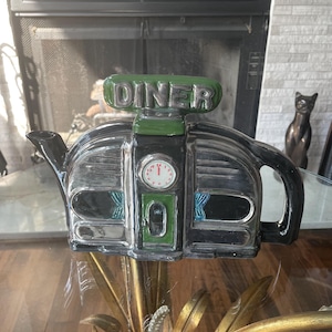 May include: A ceramic teapot shaped like a retro diner building. The exterior is black and silver with a green roof sign that reads DINER. It features a central door, a small clock face above the door, and blue curtains visible in the windows.