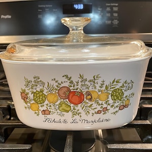 May include: White casserole dish with a glass lid. The dish has a floral design with vegetables and herbs. The text "L'Echalote La Marjolaine" is printed on the bottom of the dish.