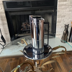 May include: A tall, chrome-finished coffee carafe with a black handle and lid sits on a matching chrome tray. The carafe is on a glass-topped table with a decorative gold base. A fireplace is in the background.