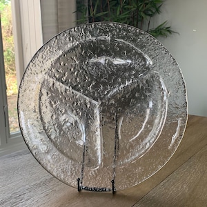 May include: A clear, textured glass plate with three divided sections. The plate is round and sits on a black metal stand. The glass has a bubbly, uneven surface. The plate is displayed on a wooden table near a window.