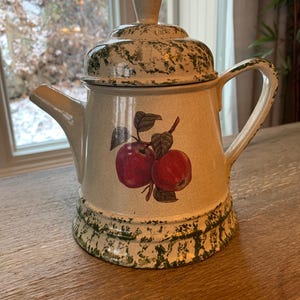 May include: A white ceramic teapot with a green and white speckled glaze. The teapot has a lid and a handle. It features a red apple design with green leaves.