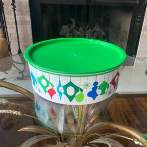 May include: A round, white container with a green lid. The container is decorated with colorful, hanging ornament designs in red, green, blue, and white. The container is sitting on a glass table.