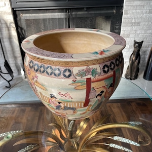 May include: A large, round ceramic fishbowl planter featuring a cream-colored base with hand-painted scenes of figures in a garden setting. The rim is decorated with intricate geometric patterns in red, blue, and gold, with a wide, open top.