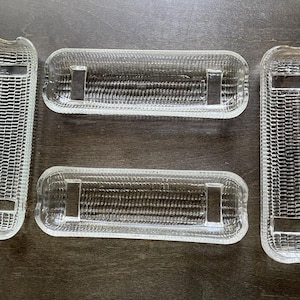 May include: Four clear, rectangular glass dishes with a textured, woven pattern. Two are long and narrow, and two are shorter and wider. Each has a rectangular indentation on each end. The dishes are arranged on a dark brown surface.